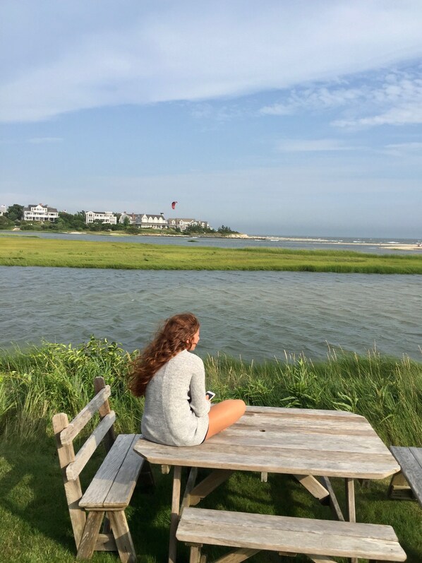 A person sitting at a wooden picnic table by a body of water with a view of a coastline and buildings in the background. - Chatham Seaside Cottage - Oceanfront Beach Cottage - Spectacular Views (Chatham)