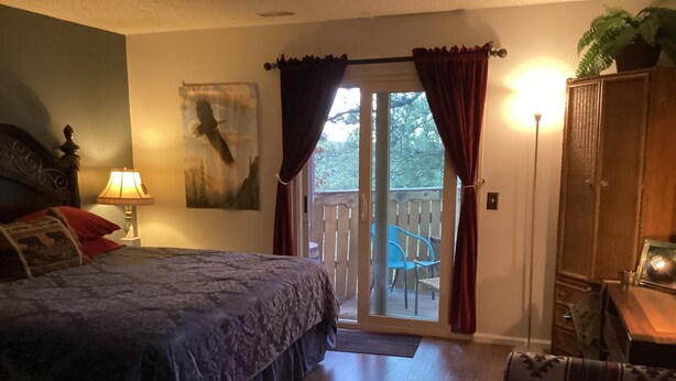 Ein Schlafzimmer mit Bett, Nachttisch, Lampe, einem Fenster mit Vorhängen, einem Balkon mit Stühlen und einem Holzschrank.