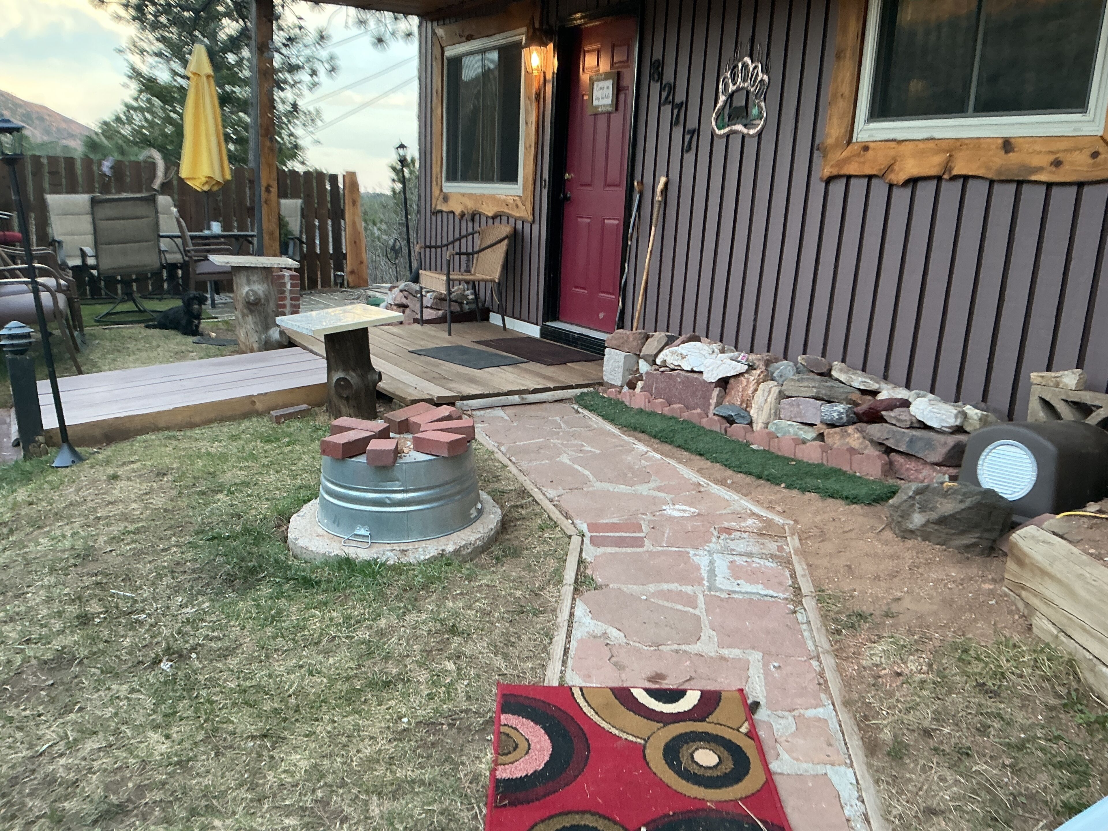 Un espace extérieur rustique avec un chemin de pierre menant à une terrasse en bois, entouré d’une clôture en bois et d’un petit jardin.