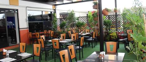 Un área de comedor al aire libre con mesas y sillas, rodeada de plantas en macetas y un suelo de césped artificial.