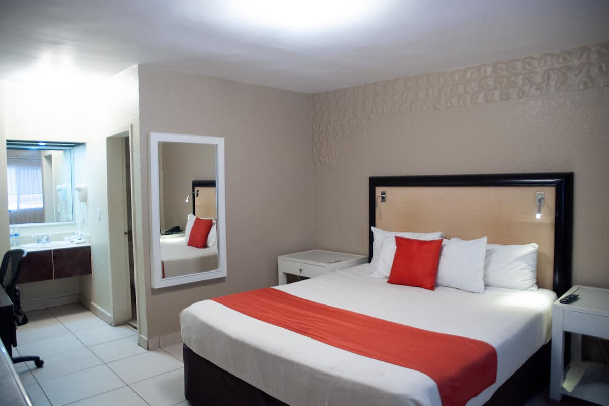 A hotel room with a large bed, a bedside table, a nightstand, a mirror, a desk with a chair, and a window with curtains.