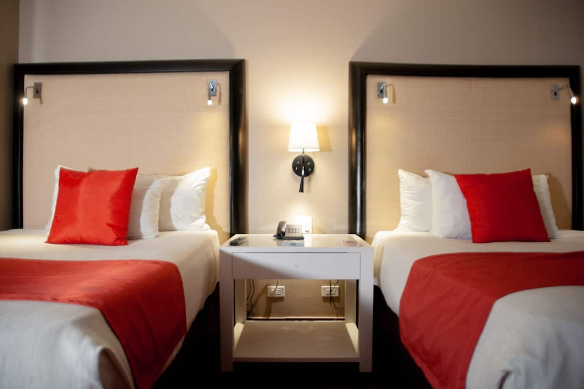 A hotel room with two beds, each with red and white bedding, a bedside table with a lamp, and a phone.