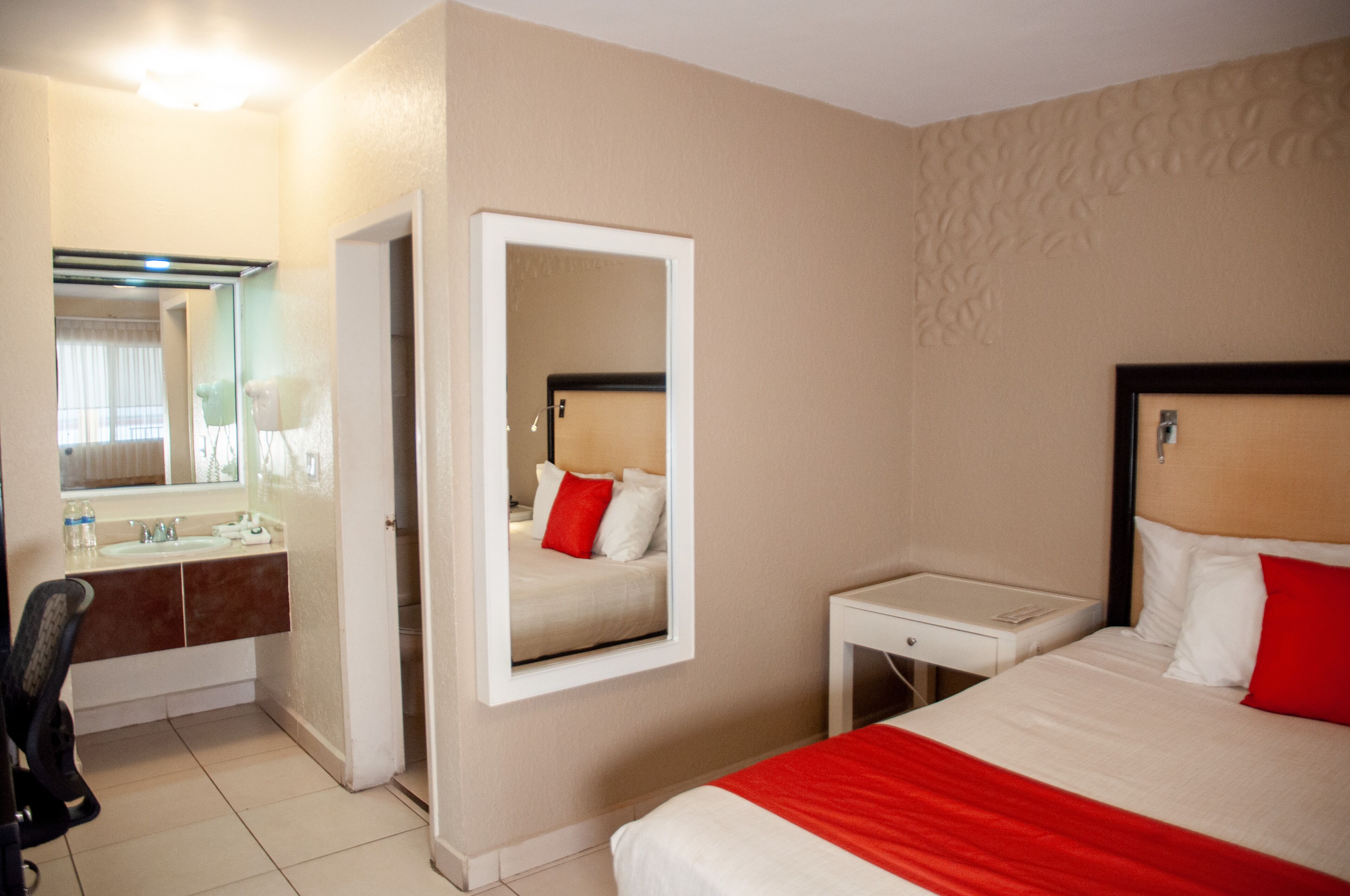 A hotel room with a bed, a bedside table, a mirror, a bathroom with a sink and mirror, and a chair.