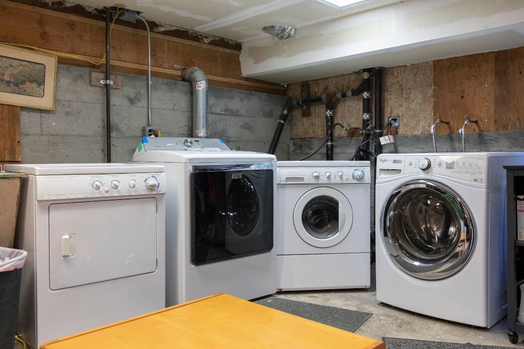 Een wasserette met drie wasmachines, een houten tafel en een betonnen muur met blootliggende leidingen.