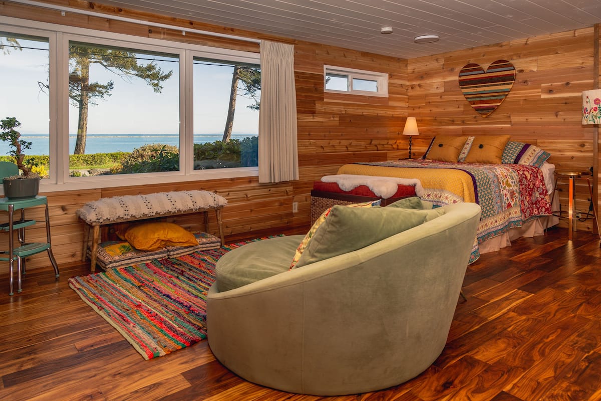 A cozy cabin with a large window, a bed, a bean bag chair, and a small table.