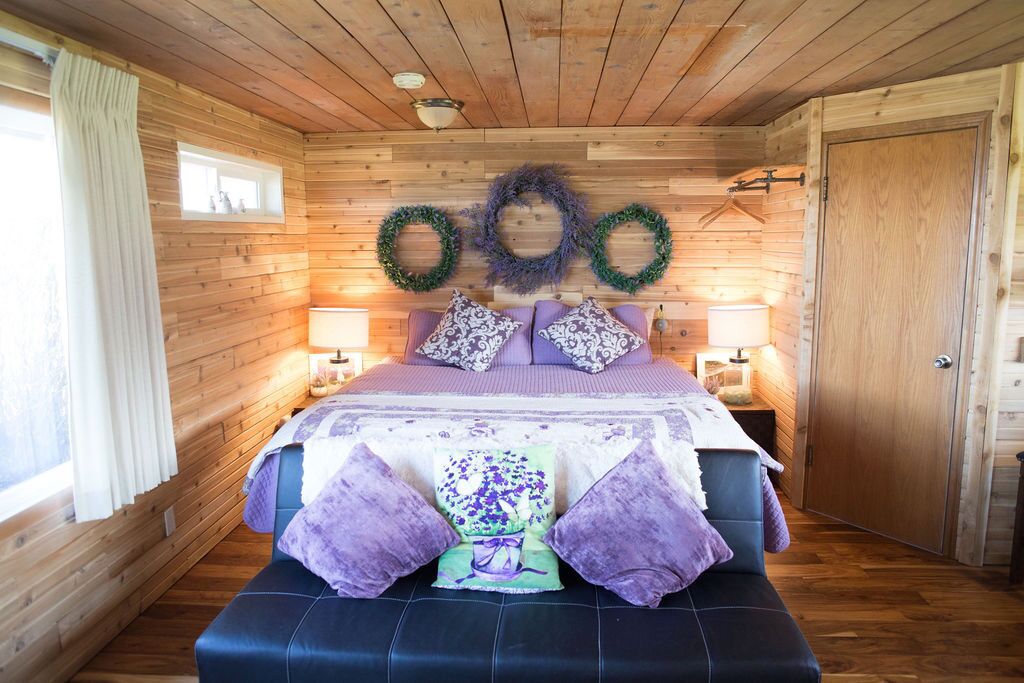 A wooden cabin bedroom with a bed, two bedside lamps, a window with curtains, and a door.