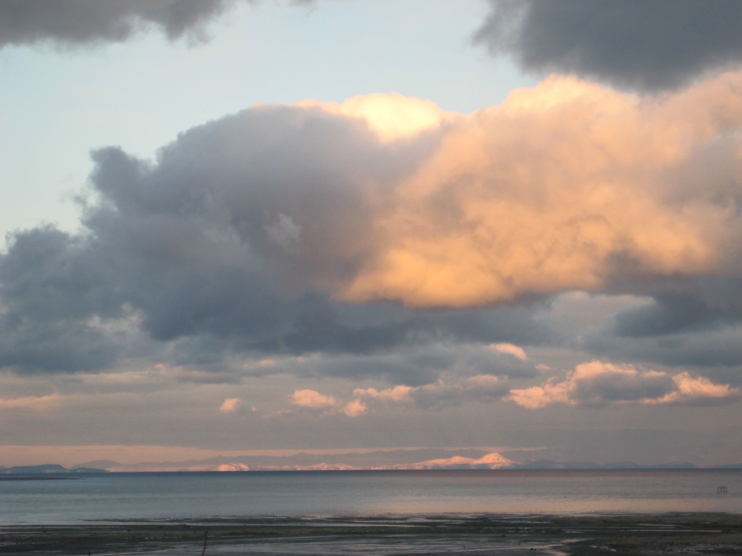 A coastal landscape with a view of the sea and distant mountains under a cloudy sky.