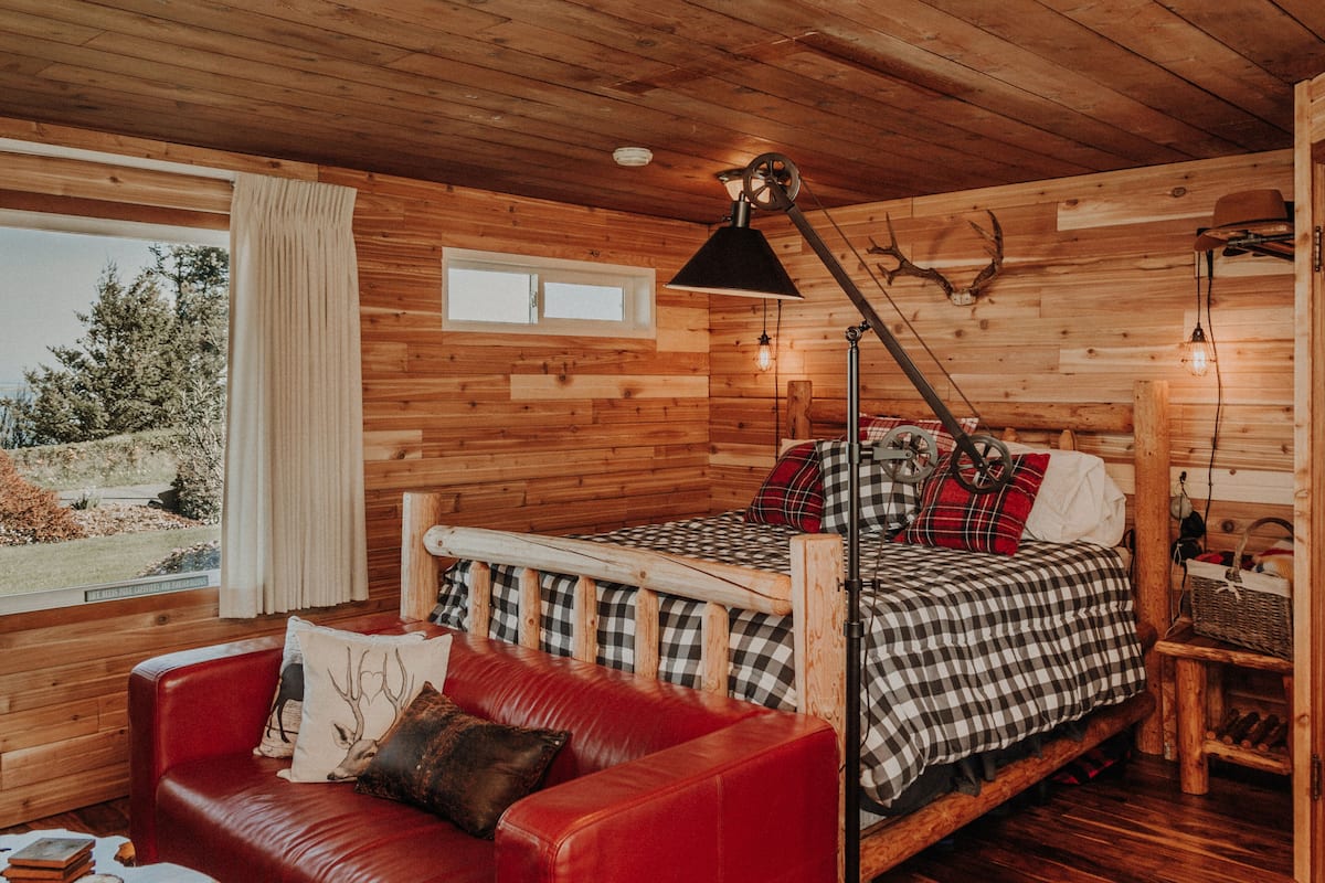 A wooden cabin room with a bed, a red leather couch, and a window overlooking a forest.