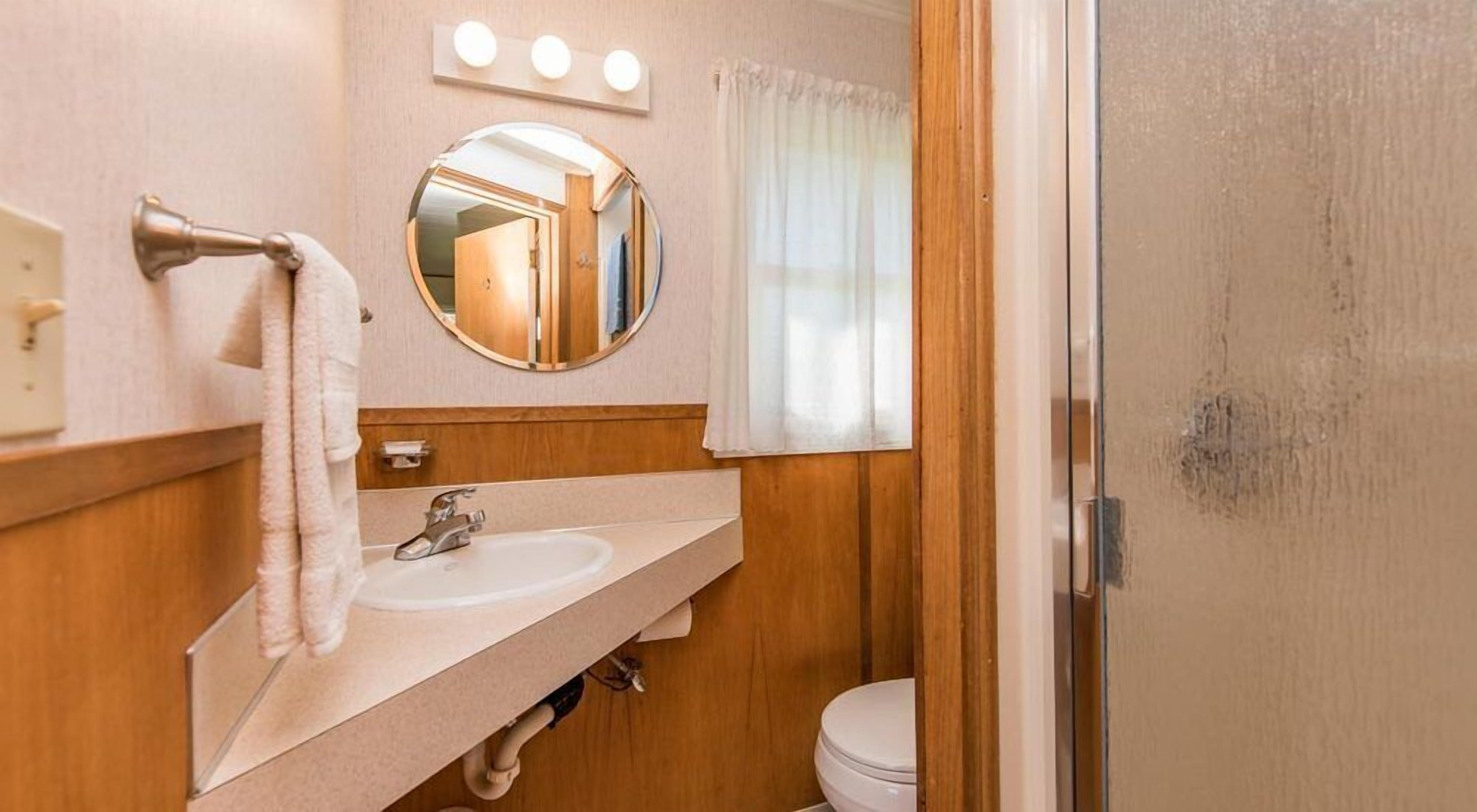 Un baño con tocador de madera, un espejo redondo, un lavamanos y un inodoro.