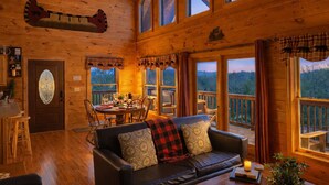A cozy cabin interior with a dining area, a sofa, and large windows offering a view of the forest. - ♡♡Mtn View W/easy Access, Hot Tub, WiFi,  Smart TV & 10 Minutes to Attractions♡♡ (Pigeon Forge)