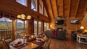 A wooden cabin interior with a dining area, fireplace, and large windows overlooking a mountainous landscape. - ♡♡Mtn View W/easy Access, Hot Tub, WiFi,  Smart TV & 10 Minutes to Attractions♡♡ (Pigeon Forge)