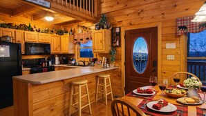 A cozy cabin-style kitchen with wooden cabinets, a central island with stools, a dining area with a table set for two, and a large window with a view of the outdoors. - ♡♡Mtn View W/easy Access, Hot Tub, WiFi,  Smart TV & 10 Minutes to Attractions♡♡ (Pigeon Forge)
