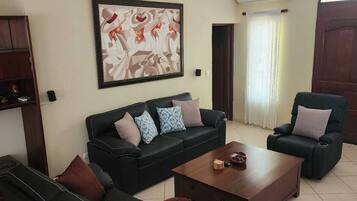 Un salon avec un canapé noir, une table basse en bois et un fauteuil vert. Un tableau est accroché au mur, et un appareil de climatisation est fixé au mur.