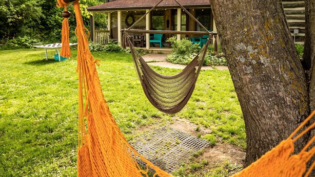 Eine Hängematte, die zwischen zwei Bäumen in einem Garten gespannt ist, mit einem Holzhaus im Hintergrund.