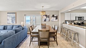 A modern living room with a blue sofa, a dining area with a wooden table and chairs, and a kitchen with white cabinets and a stainless steel oven. - Amazing 8th Floor Condo, Clean, Sleeps 10, Near Main St. | AshWorth - 801 (North Myrtle Beach)