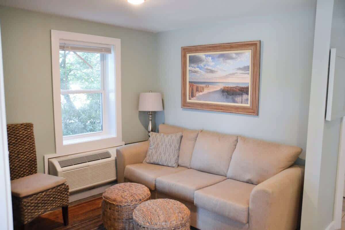 A living room with a beige sofa, two wicker ottomans, a chair, a window, a wall-mounted lamp, and a framed picture.