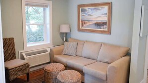 A living room with a beige sofa, two wicker ottomans, a chair, a window, a wall-mounted lamp, and a framed picture. - Steps to Beach 2BR 2BA duplex (Atlantic Beach)