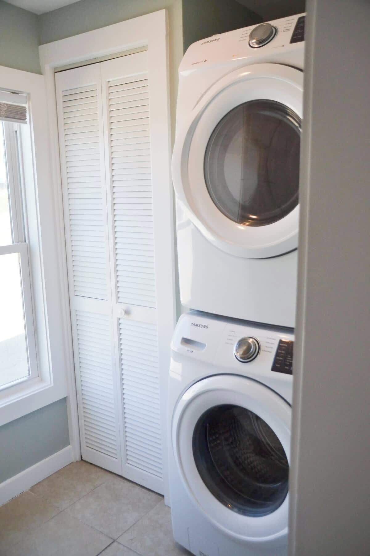 A laundry room with two stacked front-loading washing machines, a white door with louvered panels, and a window to the left.