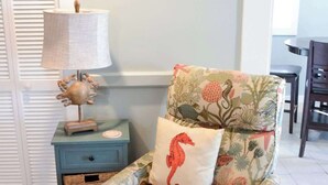 A chair with a floral pattern and a seahorse pillow, a lamp, a small table, and a wicker basket. - Steps to Beach 2BR 2BA duplex (Atlantic Beach)