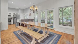 A spacious dining area with a wooden table and benches, a chandelier, and large windows offering a view of the outdoors. - Award-Winning Resort Amenities | Grand Pavilion 76 (Isle of Palms)