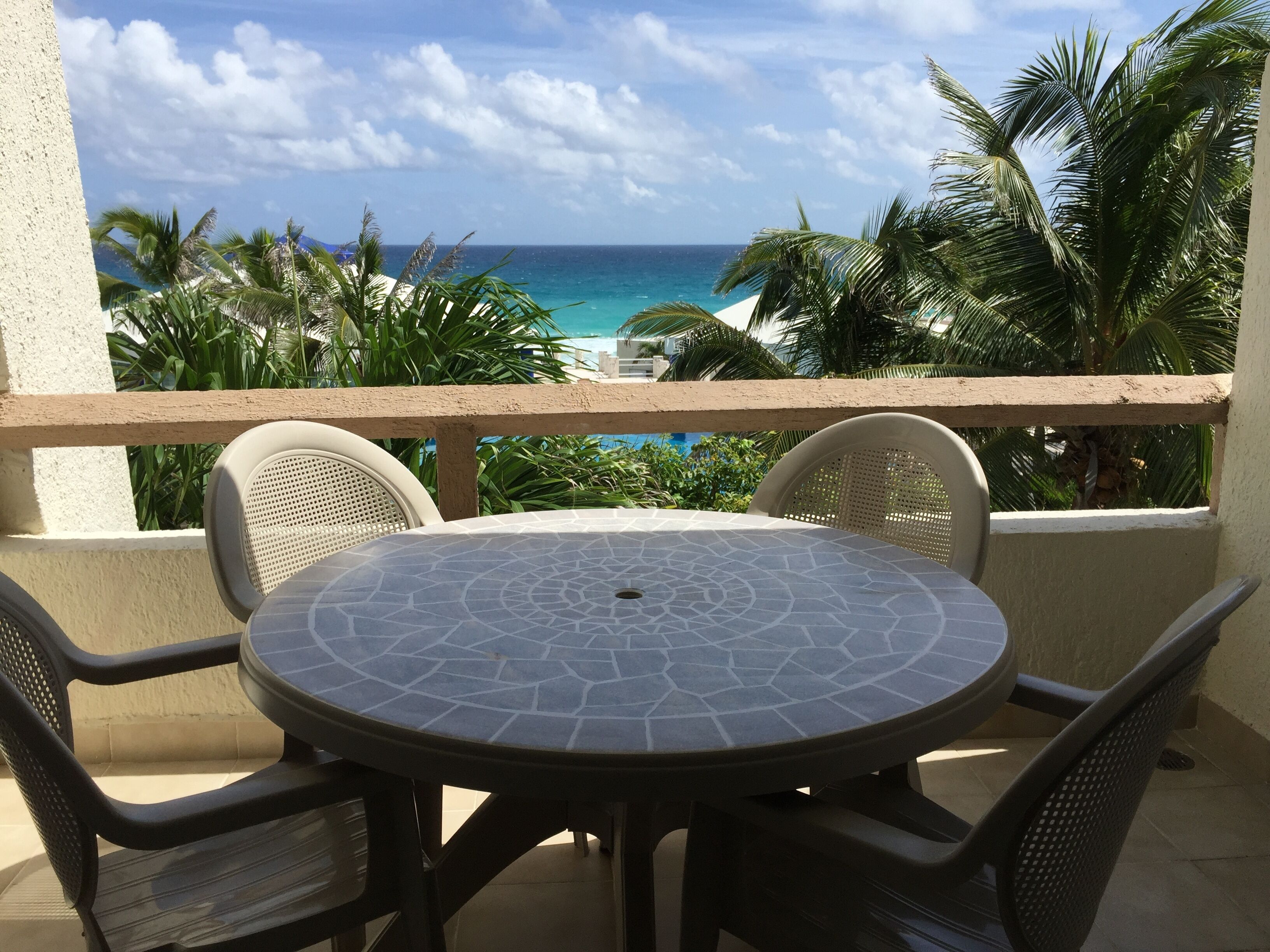 Une table ronde surmontée d’un plateau en mosaïque, entourée de quatre chaises en osier, donnant sur une plage bordée de palmiers et sur l’océan.