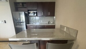 A kitchen with dark cabinets, a stainless steel refrigerator, a microwave, and a granite countertop. There are two white chairs under the counter. - Affordable Cancun Oceanfront Condo at the "Zona Hotelera" Hotel Zone (Cancun)
