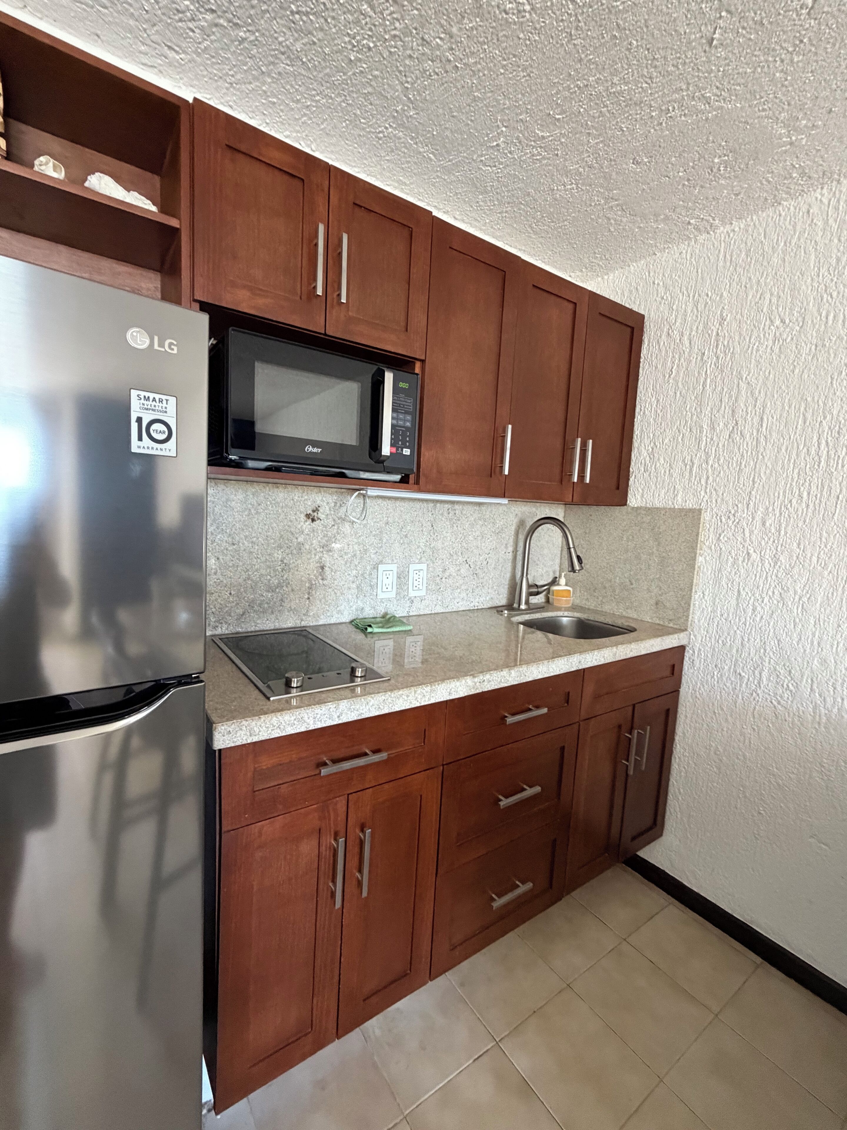 A compact kitchen with wooden cabinets, a stainless steel refrigerator, a microwave, a sink, and a countertop with a built-in stovetop.