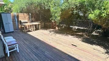 Una terraza de madera con mobiliario exterior, una parrilla para barbacoa y una vista de un edificio al fondo.