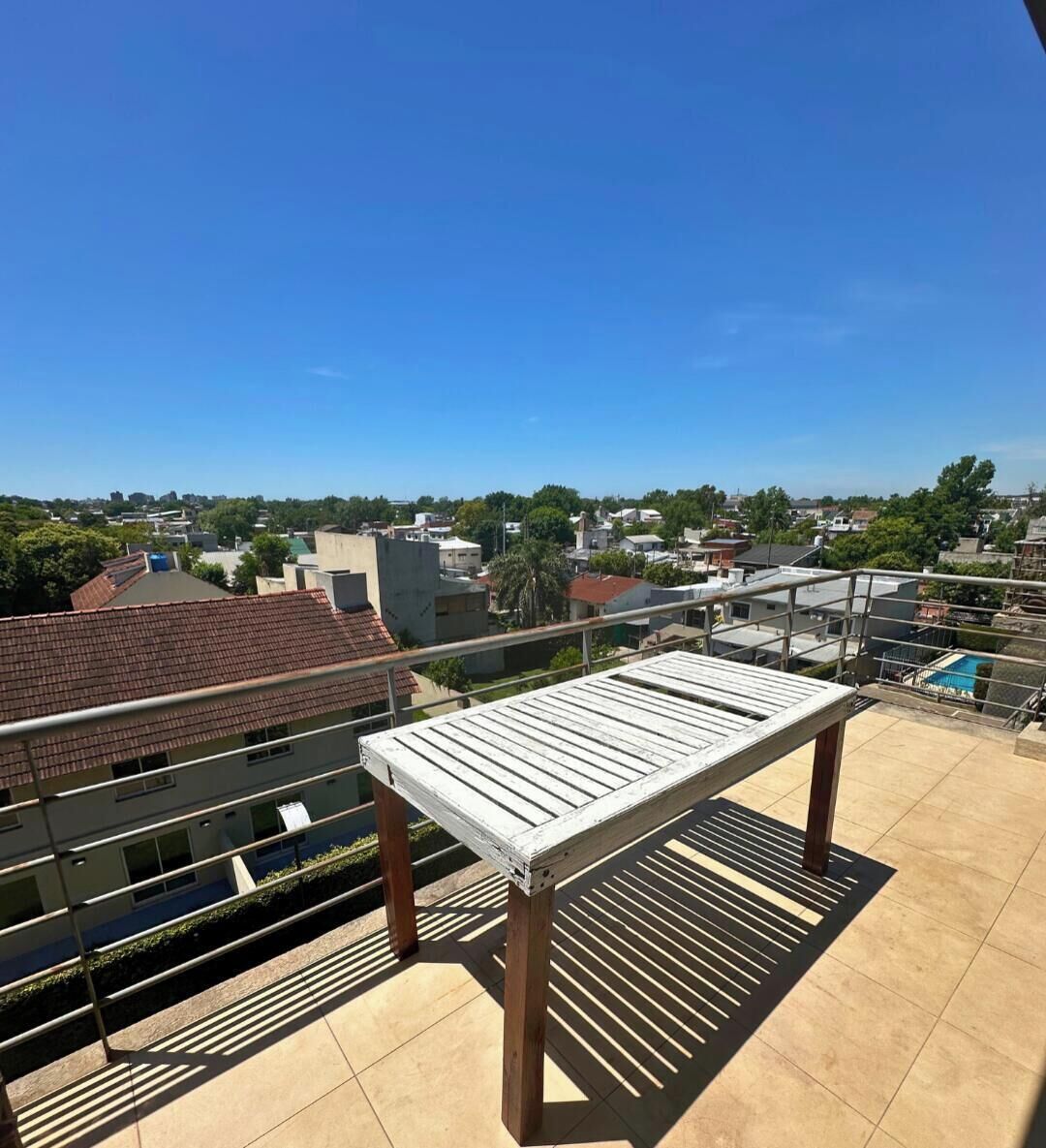 Una terraza en la azotea con barandilla metálica, un banco de madera y vistas a una zona residencial.
