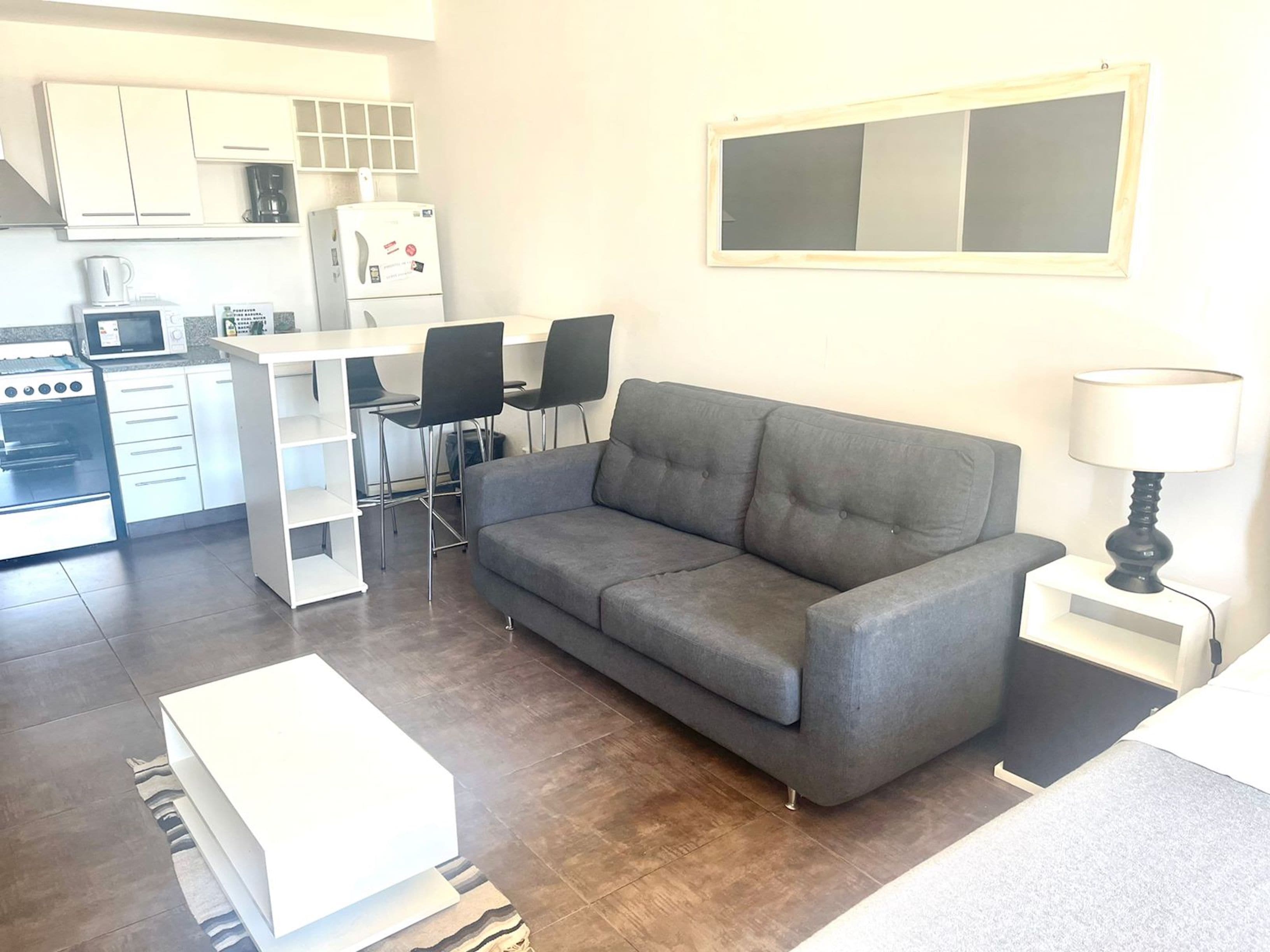 Ett kompakt bostadsutrymme med ett litet kök, en grå soffa, en matplats och ett litet bord med en lampa.