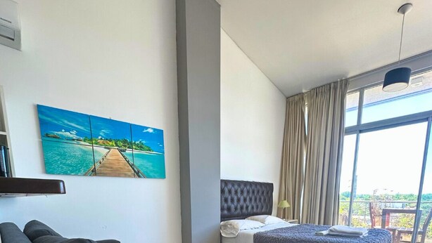 A modern hotel room with a large bed, a sofa, a painting of a tropical beach, and a balcony with a view. - Departamento Alassio (Tigre)