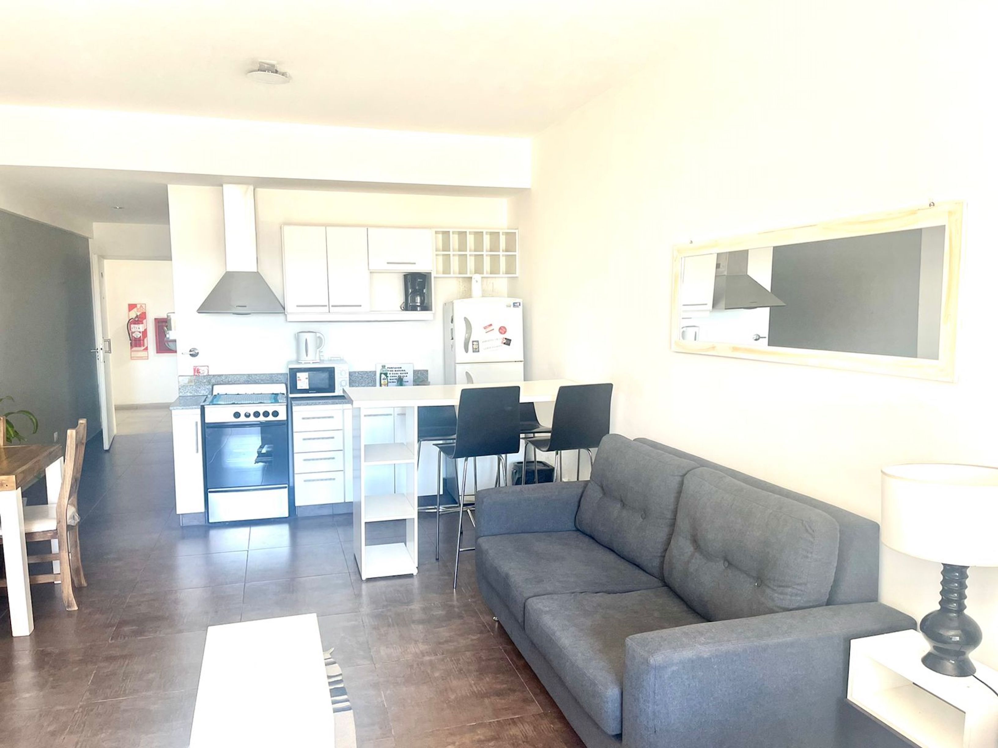 Una cocina moderna con armarios blancos, horno empotrado y microondas. Hay una zona de comedor con una mesa y sillas de madera. Un sofá gris se encuentra en la sala de estar, y una lámpara sobre una mesita.