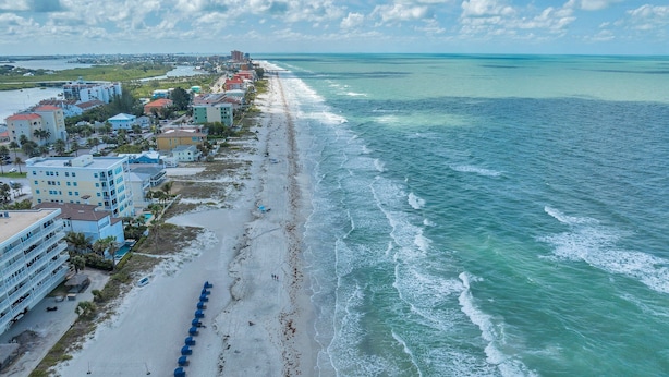 An aerial view of a coastal hotel with multiple buildings, a beach with umbrellas, and a view of the ocean.