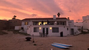 Une maison à deux étages avec un toit plat, une terrasse dotée de mobilier d’extérieur et un sol sablonneux.