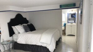 Une chambre à coucher avec un lit, une table de chevet et une chaise. On aperçoit une salle de bain à l’arrière-plan, avec un lavabo et un miroir.