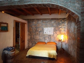 Un dormitorio con pared de piedra, una cama con ropa de cama naranja, una puerta de madera y una mesita de noche con una lámpara.