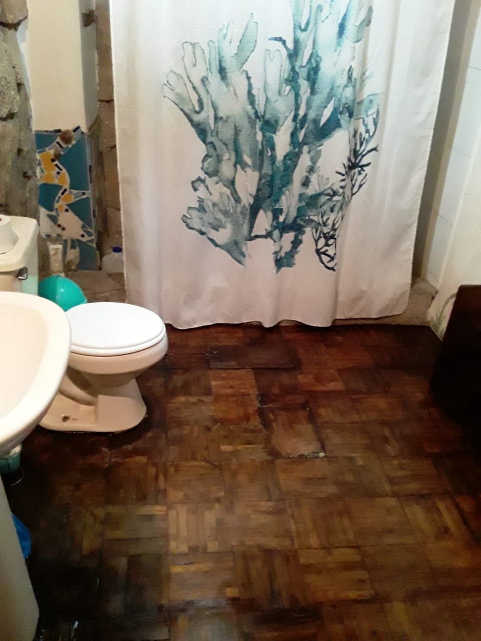 Une salle de bain avec un plancher en bois, des toilettes blanches et un rideau de douche orné d’un motif abstrait bleu.