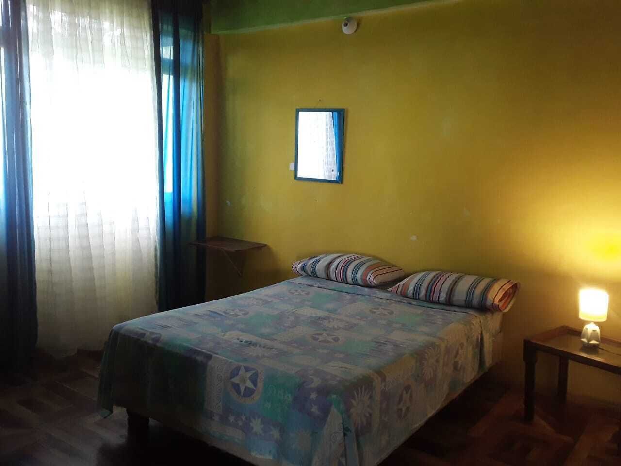 Una habitación con una cama, una mesita de noche, una lámpara de pared y un ventanal con cortinas.