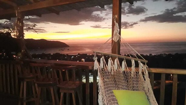 Un fauteuil suspendu avec un coussin vert, placé sur une terrasse en bois d’où l’on peut admirer un magnifique coucher de soleil.