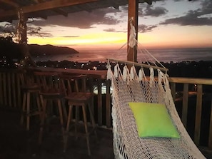 Una silla hamaca con un cojín verde sobre una terraza de madera con vista a un atardecer.