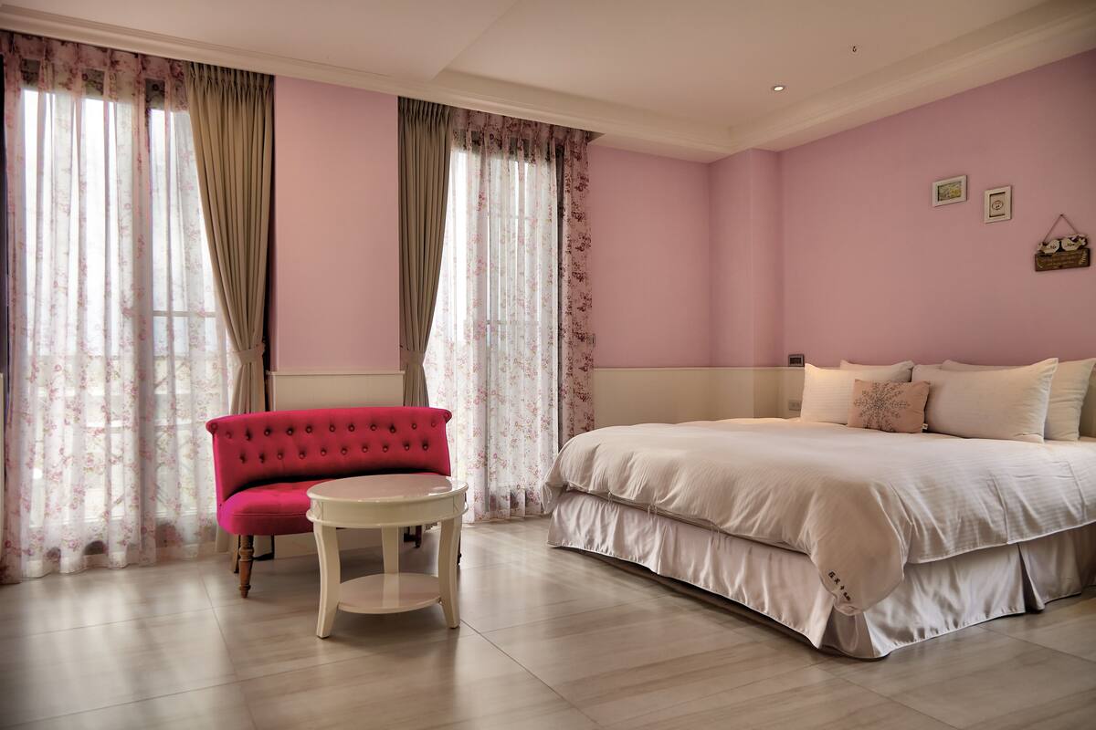 Quarto com uma cama, uma cadeira vermelha, uma mesinha redonda e cortinas.