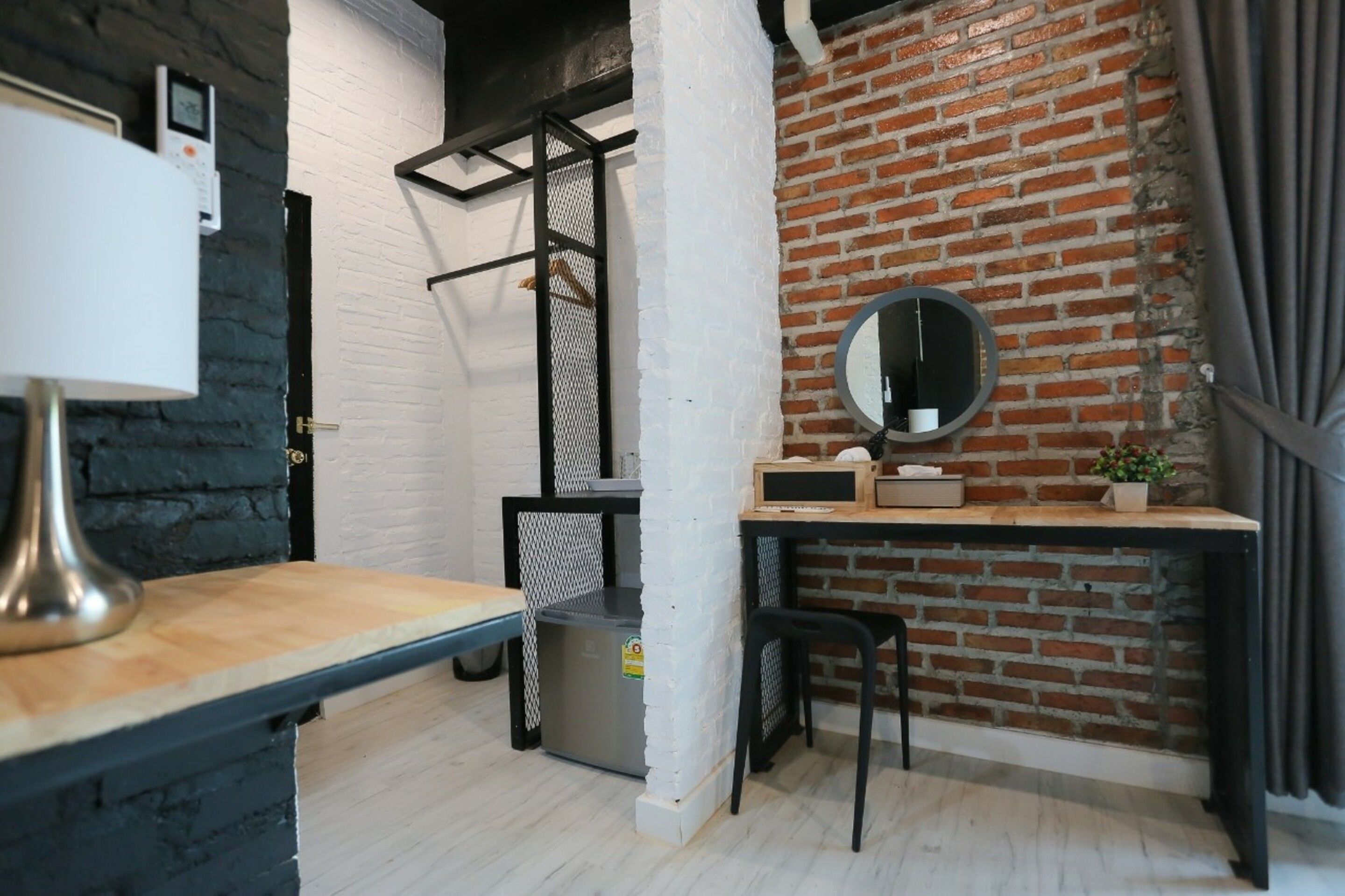 A compact room with a brick wall, a small desk with a mirror, a bench, and a trash bin.
