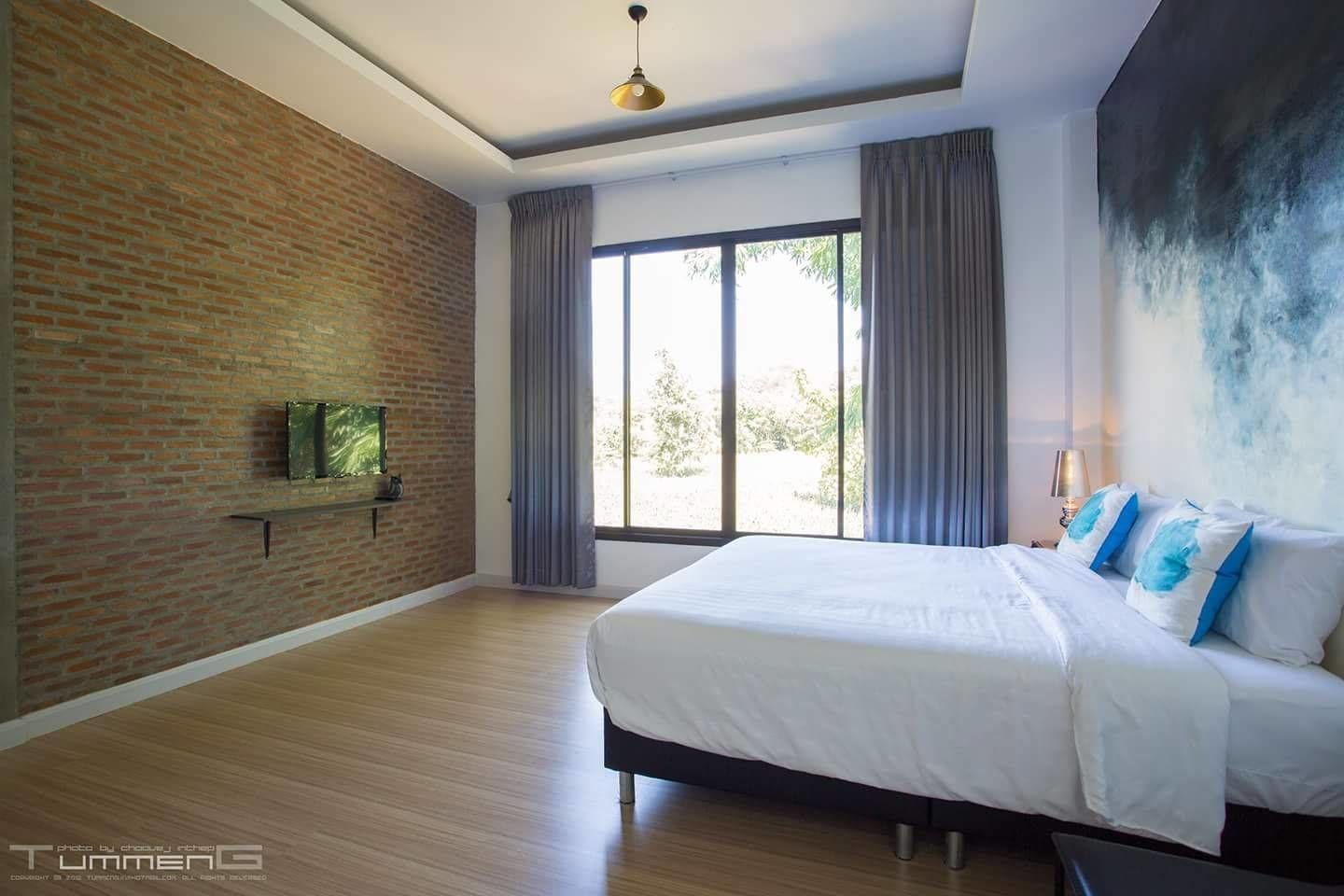 A bedroom with a brick wall, a large window with curtains, a bed with white bedding, and a mounted TV.