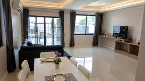 Een moderne woonkamer met een zwarte bank, een eettafel met witte stoelen en een televisie op een houten plank.