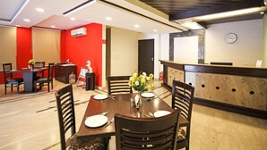 A hotel dining area with a wooden table set for four, surrounded by chairs. There is a reception desk with a clock on the wall. - Southview Apartment (New Delhi)