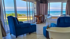 Une chambre d’hôtel dotée d’une grande porte-fenêtre coulissante donnant sur un balcon avec vue sur la mer. On y trouve un fauteuil bleu, une petite table avec une bougie et un lit blanc avec une tête de lit bleue.