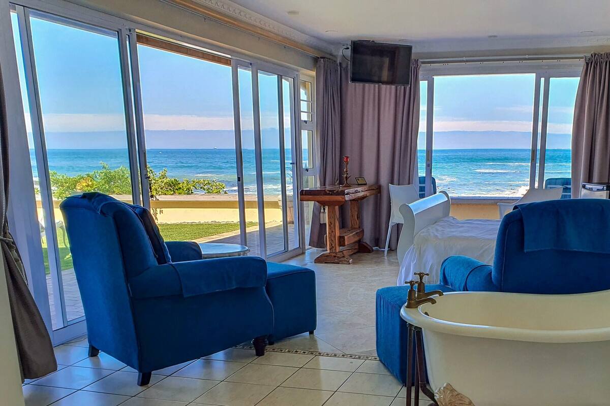 A hotel room with a large sliding glass door leading to a balcony with a view of the sea. There is a blue armchair, a small table with a candle, and a white bed with a blue headboard.