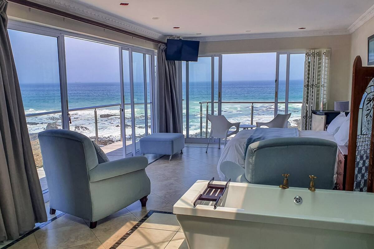 A hotel room with a large window overlooking the beach, a bed, a small table, and a chair.