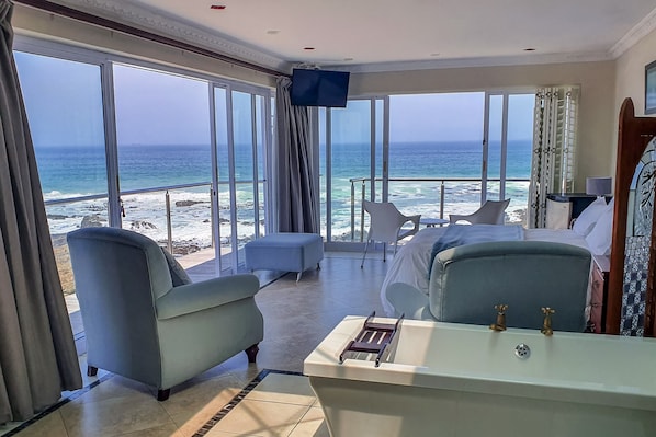 A hotel room with a large window overlooking the beach, a bed, a small table, and a chair.