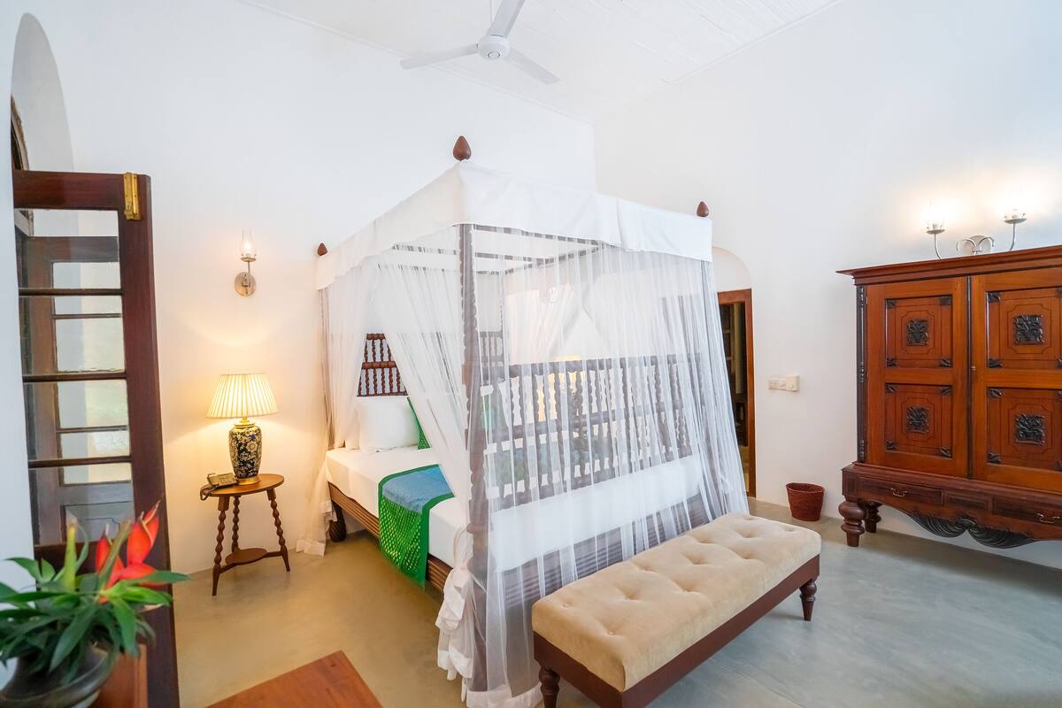 A bedroom with a four-poster bed, a bedside table with a lamp, a green throw, a wooden cabinet, and a ceiling fan.